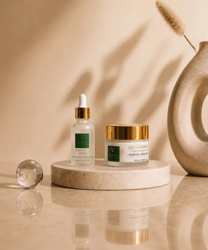 Two skincare products from the brand Vove skin: a clear glass dropper bottle labeled 'Hydro Glow Serum' and a white jar labeled 'Gluta complex' with a gold lid, displayed on a round marble pedestal with a glass sphere and a decorative beige vase in the background.